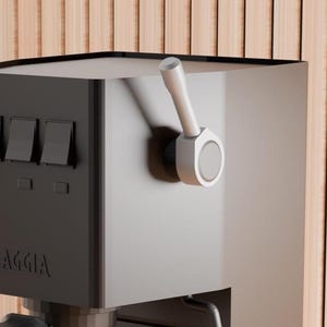 Op de afbeelding: Een zwarte espressomachine met de merknaam "GAGGIA" op de voorkant. De machine heeft een zilveren handgreep, drie zwarte schakelaars en een zwarte lekbak. De machine staat op een licht houten oppervlak.