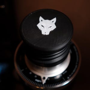 Könnte beinhalten: Schwarzer Flaschenverschluss mit weißem Wolfskopf-Design. Der Verschluss besteht aus einem dunklen Material und sitzt auf einem silbernen und schwarzen Zifferblatt. Der Wolfskopf ist ein einfaches, stilisiertes Design.
