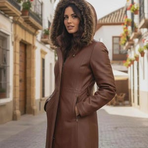 May include: A brown leather coat with a fur-trimmed hood. The long coat features a zip closure and pockets. The woman is wearing the coat outdoors.