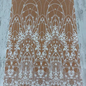 May include: A sheer fabric featuring an intricate white embroidered lace pattern with floral motifs and arched geometric lines. The design includes delicate stitching and scalloped edges, layered over a solid tan background surface.