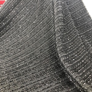 May include: Close-up of a black fabric with a diagonal striped pattern. The fabric is heavily embellished with small, shiny beads, creating a textured and shimmering effect. The beads are evenly spaced, adding to the fabric's luxurious appearance.