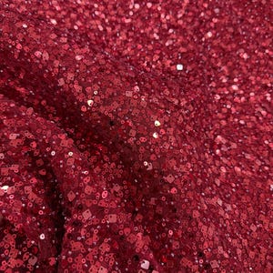 Puede incluir: Primer plano de una tela rojo oscuro cubierta de lentejuelas. Las lentejuelas son una mezcla de formas redondas y cuadradas, creando una superficie texturizada. La tela parece estar drapeada, con pliegues y sombras que añaden profundidad.