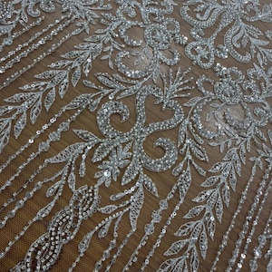 Può includere: Primo piano di tessuto in pizzo argentato con perline. Il design intricato presenta motivi floreali e a foglie con paillettes e perline scintillanti. Il pizzo è posto su un fondo a rete marrone, creando una texture delicata ed elegante.
