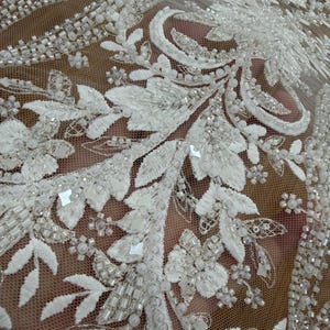 May include: Close-up of white beaded lace fabric. The lace features intricate floral and leaf patterns, embellished with beads and sequins. The delicate design is set against a sheer, net-like background, creating a textured and elegant appearance.