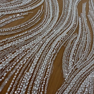 Puede incluir: Primer plano de una tela con un respaldo de malla beige, adornada con patrones ondulados de cuentas blancas y lentejuelas plateadas. Las cuentas varían en tamaño, creando un efecto texturizado y brillante. Ideal para vestidos de novia o de noche.