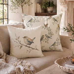 May include: Two square throw pillows with bird illustrations on a beige sofa. One pillow features a single bird on a branch, the other has multiple birds and foliage. The pillows add a touch of nature-inspired decor.