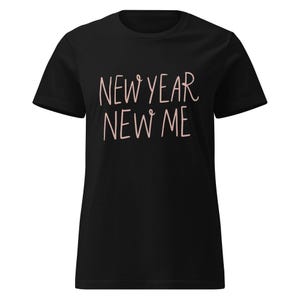 以下が含まれることがあります： 黒いTシャツ。テキスト「NEW YEAR NEW ME」は薄いピンク色の筆記体です。シャツは半袖とクルーネックです。テキストはシャツの前面の中央に配置されています。