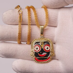 Puede incluir: Collar con colgante dorado con un colgante cuadrado con una cara negra, labios rojos y ojos blancos y rojos. El colgante está sujeto por una cadena dorada. El collar se muestra sobre un guante blanco.