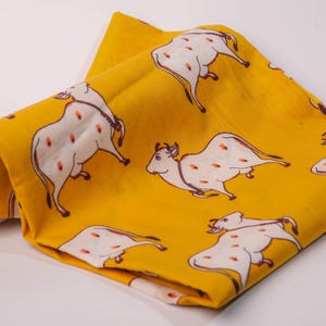 May include: A folded yellow fabric with a repeating pattern of white cows with red spots. The cows are drawn in a simple, illustrative style. The fabric is likely cotton or linen.