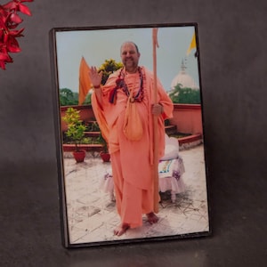 Puede incluir: Fotografía enmarcada de un hombre con túnicas naranjas, saludando. Lleva un collar de cuentas y una bolsa. El fondo muestra un edificio y árboles. El marco es negro.