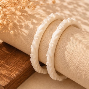 Puede incluir: Dos pulseras blancas con estampado floral descansan sobre una tela de lino beige. Las pulseras presentan un diseño repetido de flores y hojas. Un pequeño bloque de madera y flores secas están en el fondo.