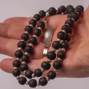 Puede incluir: Un collar de cuentas marrón oscuro con detalles plateados. El collar está compuesto por cuentas redondas de color marrón oscuro ensartadas con espaciadores plateados. El collar se sostiene en una mano.