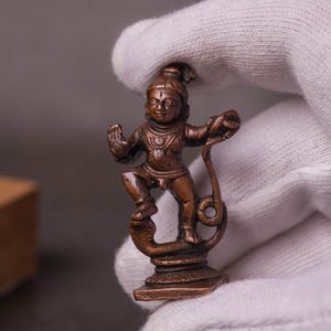 May include: A small, dark bronze-toned metal figurine depicting a deity standing on a coiled serpent. The figure has one hand raised in a gesture, wears traditional jewellery, and is mounted on a square base, showcasing intricate craftsmanship.