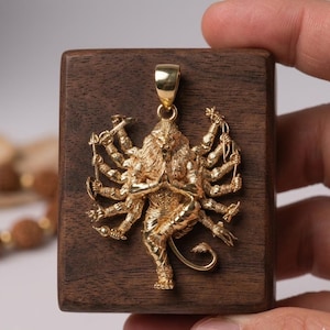 Op de afbeelding: Een goudkleurige hanger met een veelarmige, leeuwachtige figuur. De hanger is ingewikkeld gedetailleerd met meerdere armen die verschillende objecten vasthouden. De hanger is bevestigd aan een gouden beugel en is geplaatst op een donkerbruin houten blok.
