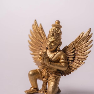 Puede incluir: Estatua dorada de una figura arrodillada con grandes alas detalladas. La figura lleva una corona y tiene las manos juntas en oración. La estatua es probablemente una representación de una figura mitológica o religiosa.