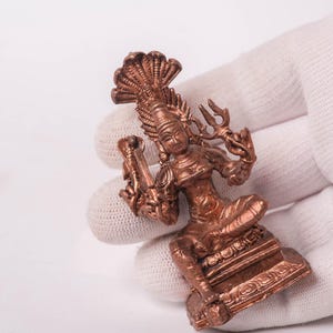 May include: A copper-coloured figurine of a deity, possibly Hindu, with a multi-headed cobra headdress and multiple arms. The figure is seated on a detailed base and holds various symbolic objects. The statue is held by a gloved hand.