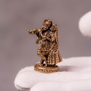 Könnte beinhalten: Eine kleine, goldfarbene Metallfigur, die zwei Figuren auf einem runden Sockel darstellt. Eine Figur spielt eine Flöte, während die andere daneben steht. Die Skulptur weist detaillierte Verzierungen an der Kleidung und den Accessoires auf.