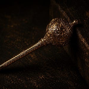 May include: A metallic gold-toned mace featuring an intricate, embossed floral and scrollwork pattern covering the entire surface of the spherical head and the elongated, slender handle, ending in a small, rounded finial tip at the base.