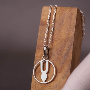 May include: A silver-toned pendant necklace featuring a circular frame enclosing a vertical U-shaped symbol with a pointed base. The pendant hangs from a delicate silver-toned chain, showcasing a polished metallic finish and a clean, symmetrical design.