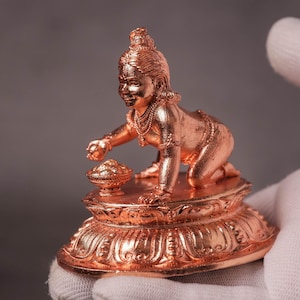 May include: A copper-colored metallic figurine of a crawling child deity positioned on a circular lotus-shaped base. The figure wears multiple necklaces and armbands, reaching toward a small bowl placed on the platform in front of them.