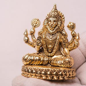 May include: A metallic gold-colored figurine depicting a deity with a lion head and human body, seated in a cross-legged position on a lotus-shaped base. The figure has four arms, holding various symbolic objects, and wears ornate jewelry.