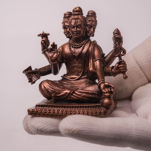 Könnte beinhalten: Eine bronzefarbene Statue einer Gottheit mit drei sichtbaren Gesichtern und mehreren Armen, die in meditativer Haltung sitzt. Die Statue ist mit Halsketten geschmückt und hält verschiedene symbolische Objekte. Der Sockel weist aufwendige dekorative Muster auf.
