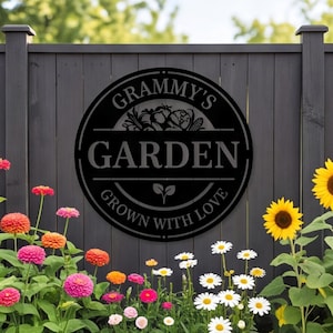 May include: A circular black metal sign featuring a cutout design with the text "GRAMMY'S GARDEN" and "GROWN WITH LOVE" along with a graphic of vegetables and a small sprout. The sign is mounted on a dark gray vertical wooden fence panel.