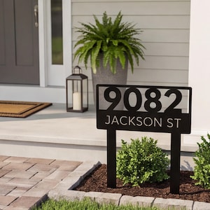 May include: A black metal yard sign featuring the house number 9082 and the text JACKSON ST. The rectangular sign is supported by two vertical stakes inserted into the ground, displaying bold, cut-out lettering and numerals in a clean font.