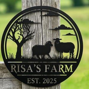 Puede incluir: Letrero de granja de metal negro con diseño circular. El letrero presenta dos ovejas, un árbol y el texto "RISA'S FARM EST. 2025". El letrero está montado en un poste de madera desgastada con un fondo verde borroso.