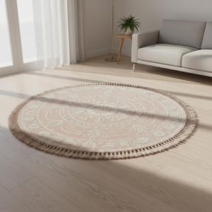 May include: A round, blush-colored rug with a detailed floral pattern in cream. The rug has a fringe border and is placed on a light wood floor. The rug is a decorative home accent.