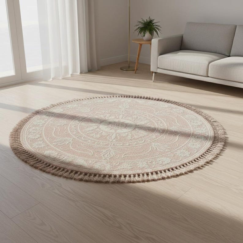 May include: A round, blush-colored rug with a detailed floral pattern in cream. The rug has a fringe border and is placed on a light wood floor. The rug is a decorative home accent.