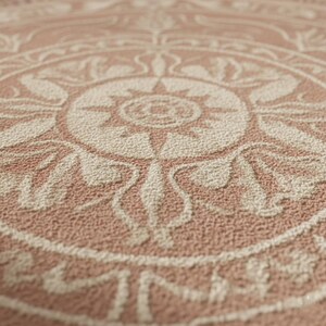 May include: Close-up of a textured, circular rug with a detailed, symmetrical design. The rug features a repeating pattern in shades of beige and dusty rose, creating a vintage or bohemian aesthetic. The design includes floral and geometric elements.