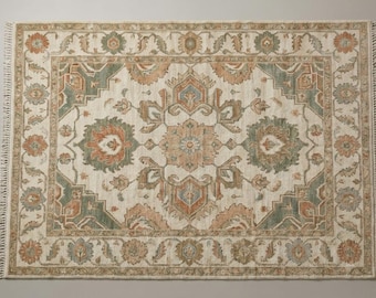 May include: A rectangular area rug featuring a cream-colored base with a symmetrical pattern in muted shades of sage green, terracotta orange, and soft blue. The design includes floral motifs and geometric borders with short fringe on the ends.