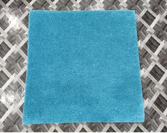 May include: A square, turquoise rug with a plush, textured surface. The rug is a solid color and appears to be made of a soft material. The rug is placed on a patterned surface.