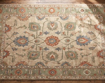 May include: A rectangular area rug featuring a beige background with a symmetrical floral pattern in shades of sage green, burnt orange, and slate blue. The rug has a wide decorative border with repeating floral motifs and short fringe edges.