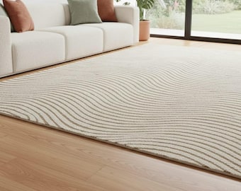 May include: A beige area rug with a wavy striped pattern. The rug is placed on a hardwood floor in front of a white sofa with accent pillows. The rug's design creates a sense of movement and texture in the room.