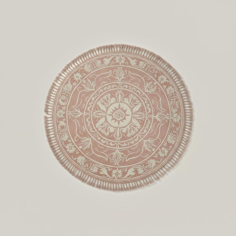 May include: A round, pink rug with a detailed floral and geometric pattern in off-white. The rug has a fringed edge and a central medallion design. The rug is a decorative home accent.