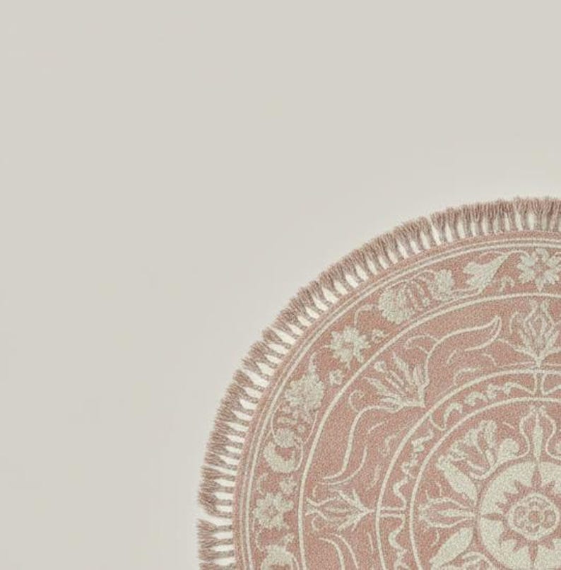 May include: A portion of a round, pink rug with a detailed floral pattern and fringe trim. The rug's design features a central medallion surrounded by stylized flowers and leaves in a lighter shade of pink.