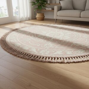May include: A round rug with a pink and cream floral design. The rug has a fringe border and is placed on a hardwood floor. The rug is in a living room setting with a couch and a potted plant visible in the background.