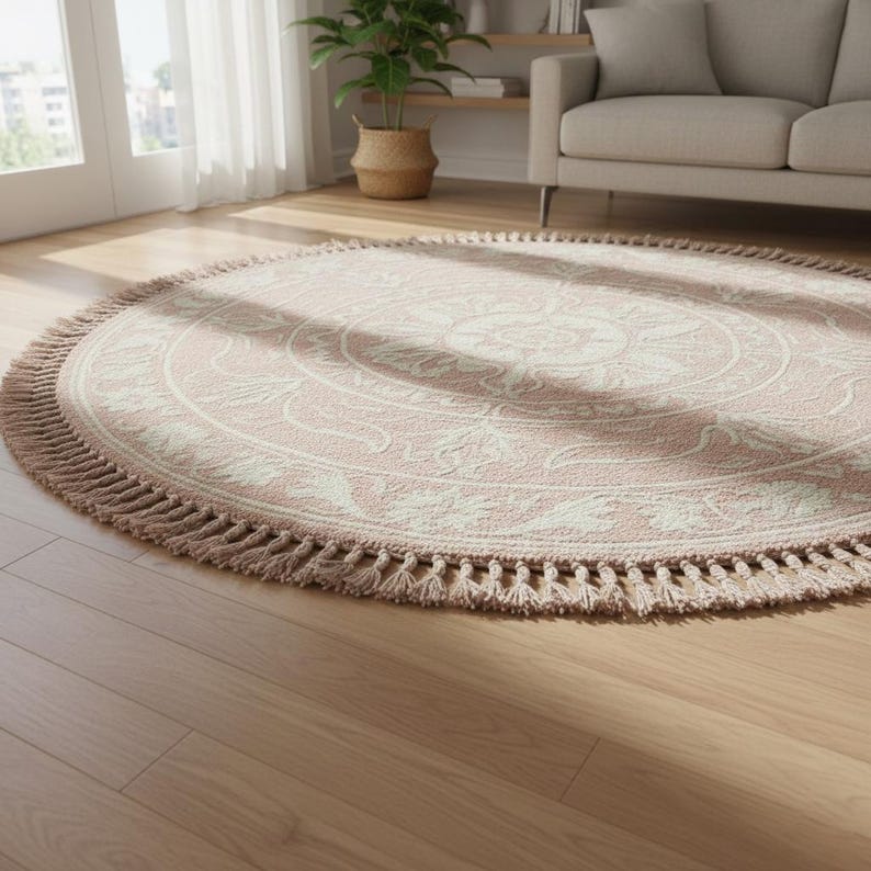 May include: A round rug with a pink and cream floral design. The rug has a fringe border and is placed on a hardwood floor. The rug is in a living room setting with a couch and a potted plant visible in the background.