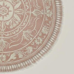 May include: A round, pink rug with a detailed floral and scrollwork design in off-white. The rug has a fringed edge and a textured appearance, suitable for home decor.