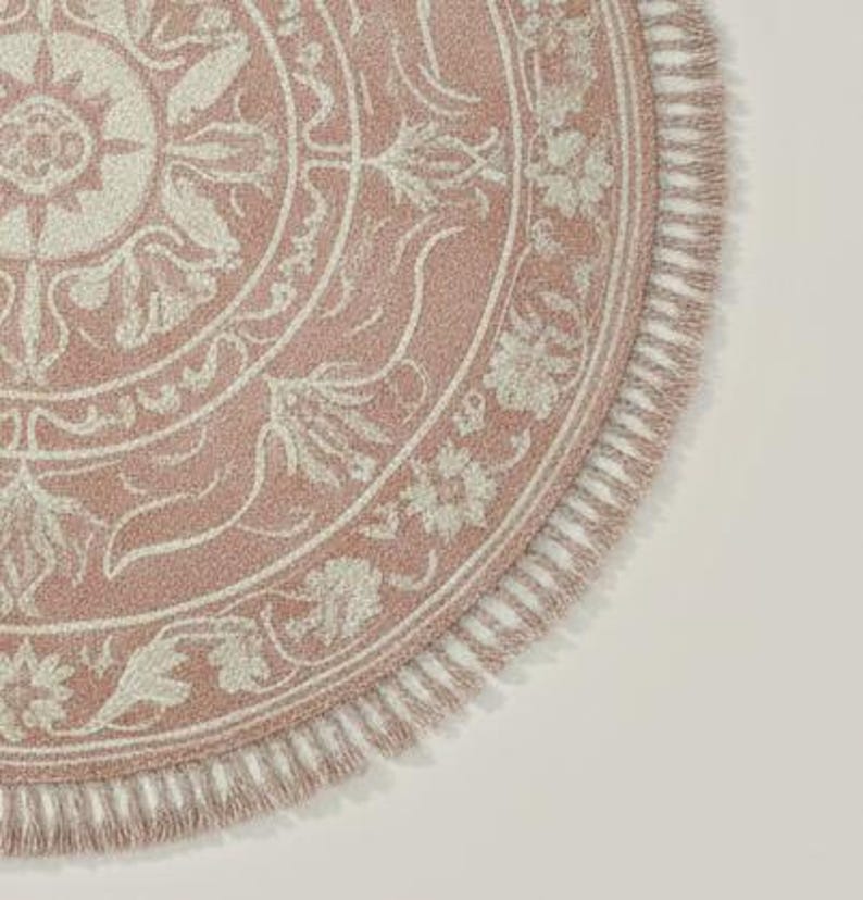 May include: A round, pink rug with a detailed floral and scrollwork design in off-white. The rug has a fringed edge and a textured appearance, suitable for home decor.
