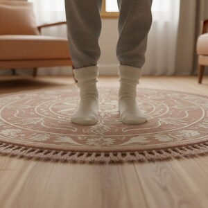 May include: A round, blush-colored rug with a detailed floral and geometric pattern. The rug has a fringe border and is placed on a wooden floor. The rug is approximately 4 feet in diameter.