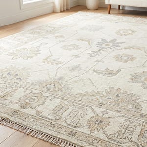 May include: A large rectangular area rug featuring an off-white base with a repeating pattern of floral and geometric motifs in muted shades of beige, light grey, and soft blue. The rug has a wide decorative border and short fringe on the edges.