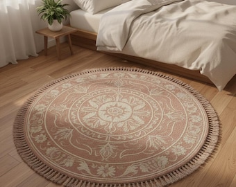 May include: A round, blush-colored rug with a cream-colored floral and geometric pattern. The rug has a fringe border and is placed on a hardwood floor. The rug is approximately 60 inches in diameter, suitable for a bedroom or living room.