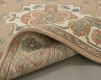 May include: Beige area rug with a detailed floral and geometric pattern. The rug features a rolled-up edge, revealing the intricate design. Colors include beige, blue, orange, and green. The rug's design is reminiscent of traditional oriental rugs.