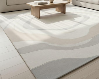 May include: A rectangular area rug with a wave pattern in shades of gray, beige, and white. The rug is placed on a light-colored wooden floor. The rug's design features soft, flowing lines, creating a sense of movement and texture.