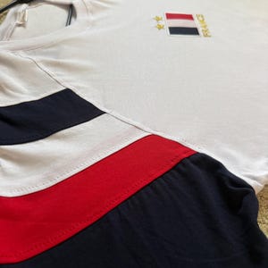 May include: A white short-sleeved t-shirt featuring navy blue and red horizontal stripes on the sleeves and chest. The chest displays an embroidered rectangular French flag patch with two gold stars above it and the word FRANCE written vertically.