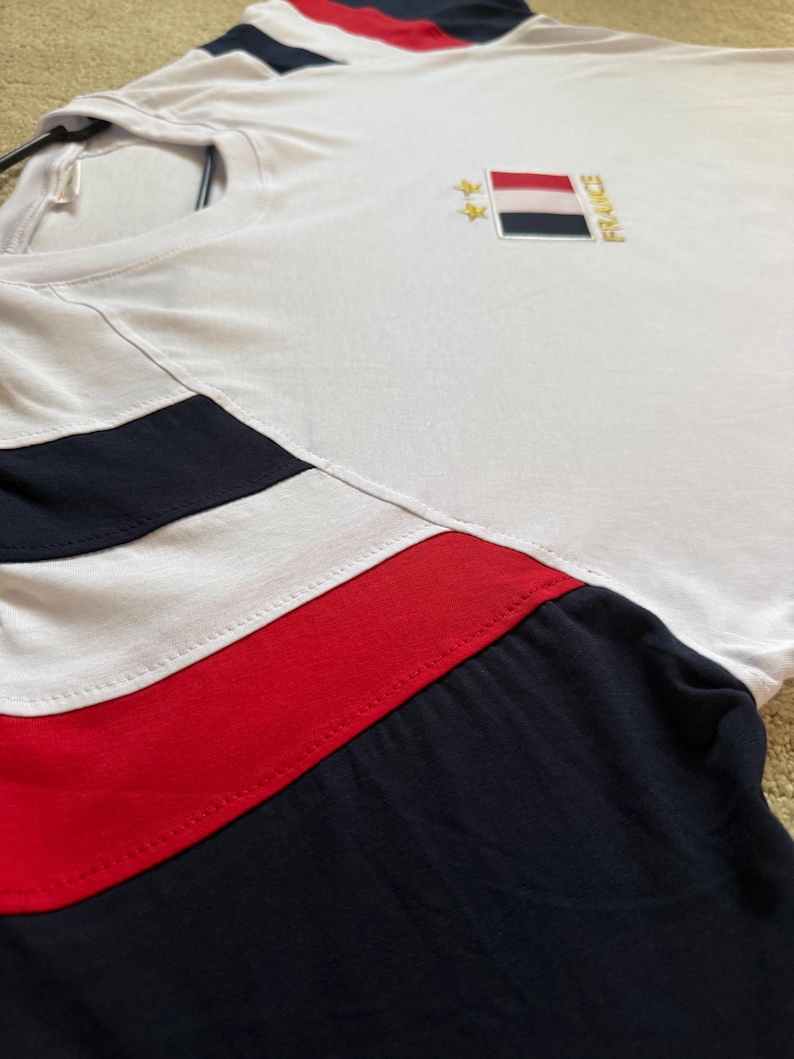 May include: A white short-sleeved t-shirt featuring navy blue and red horizontal stripes on the sleeves and chest. The chest displays an embroidered rectangular French flag patch with two gold stars above it and the word FRANCE written vertically.