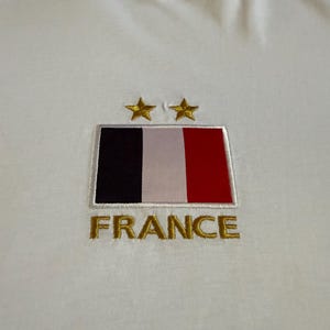 May include: A white t-shirt featuring an embroidered rectangular patch of the French flag with vertical black, white, and red stripes. Above the flag are two gold embroidered stars, and below the flag is the word FRANCE in gold embroidery.